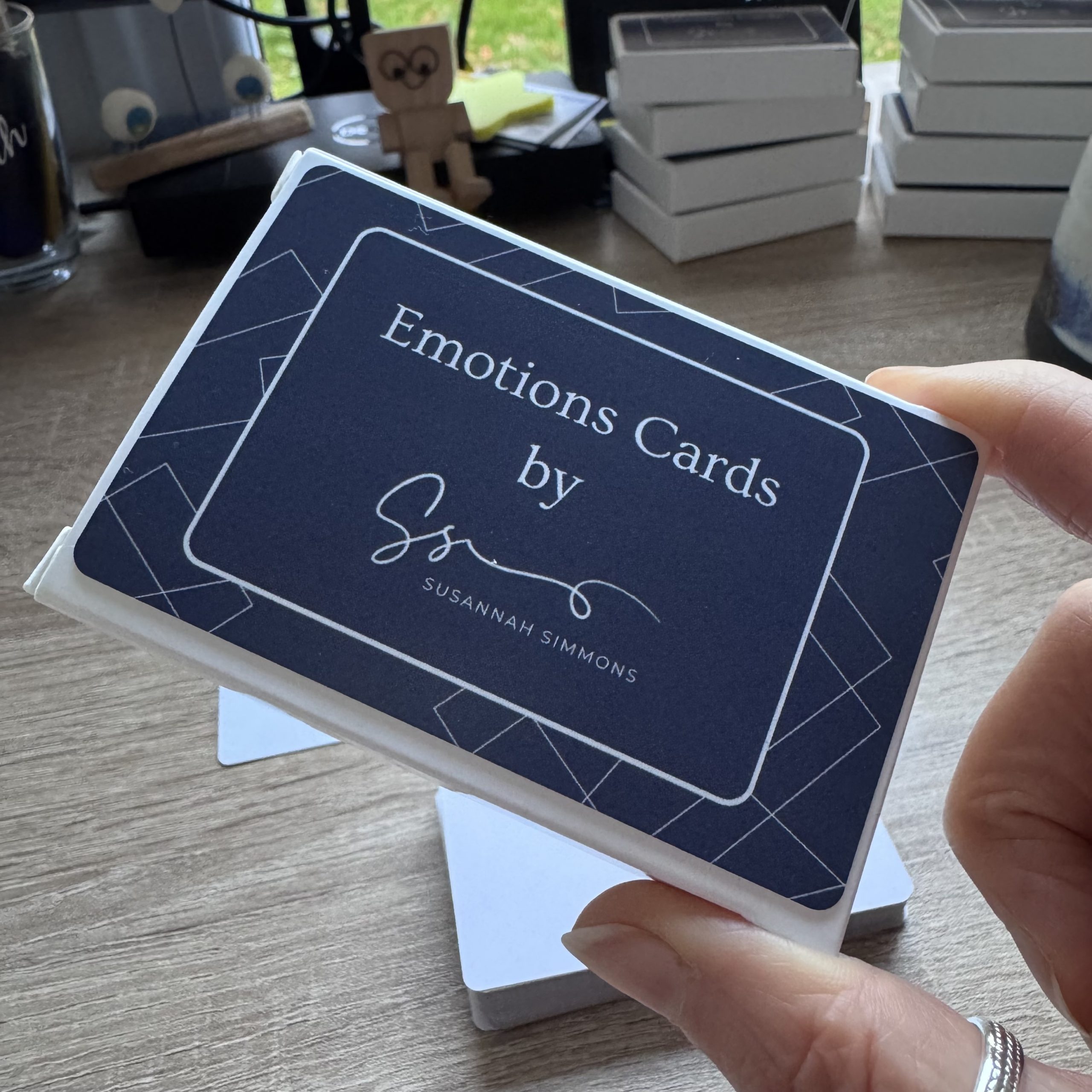 A hand holding a box of Emotions Cards by Susannah Simmons, with several other card decks stacked on a desk in the background